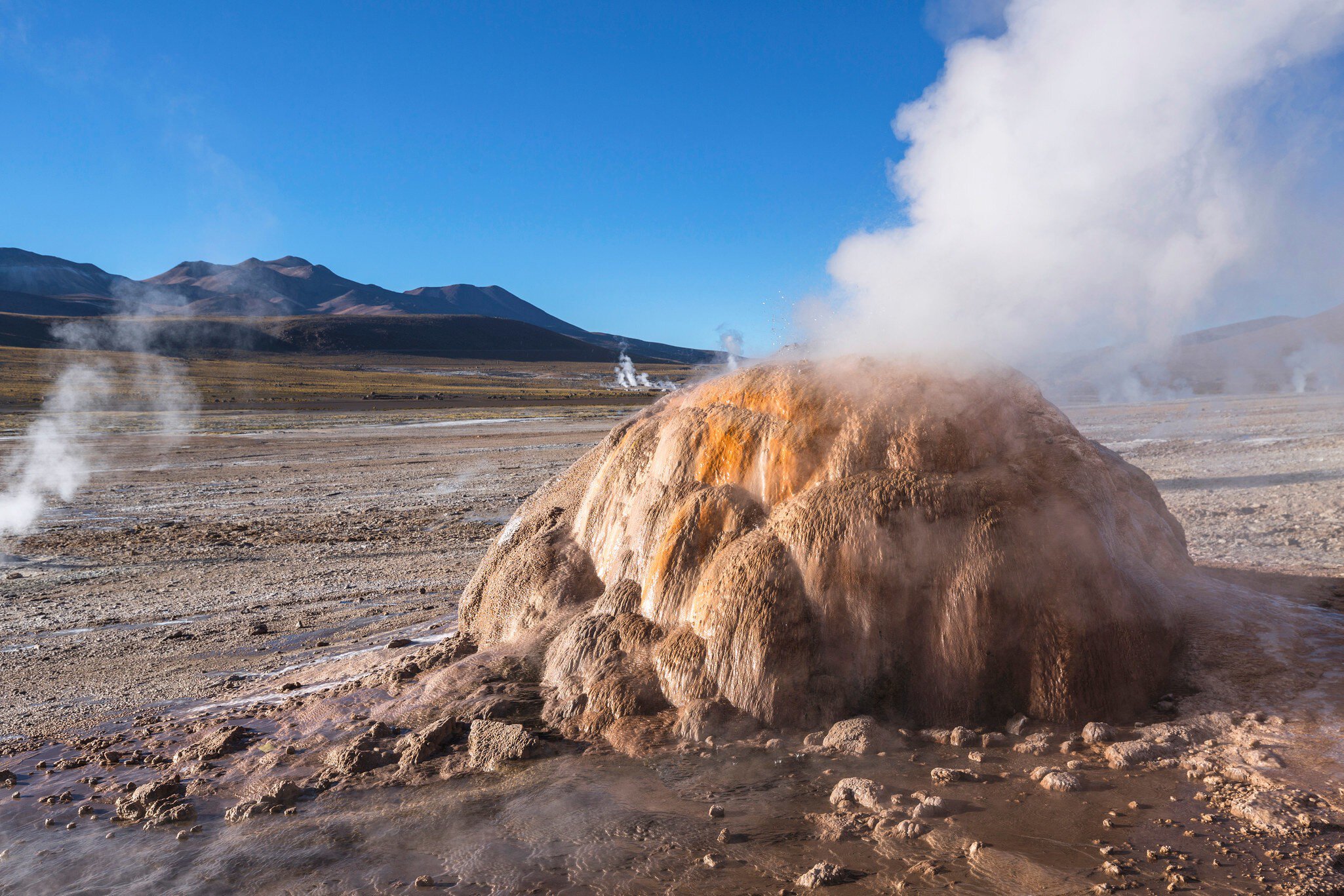 White steam billows from a rounded orange and tan mineral mound on a flat plain below brown mountains