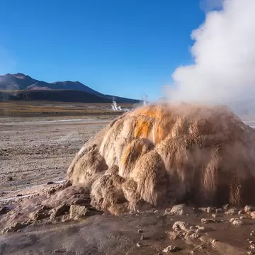 White steam billows from a rounded orange and tan mineral mound on a flat plain below brown mountains
