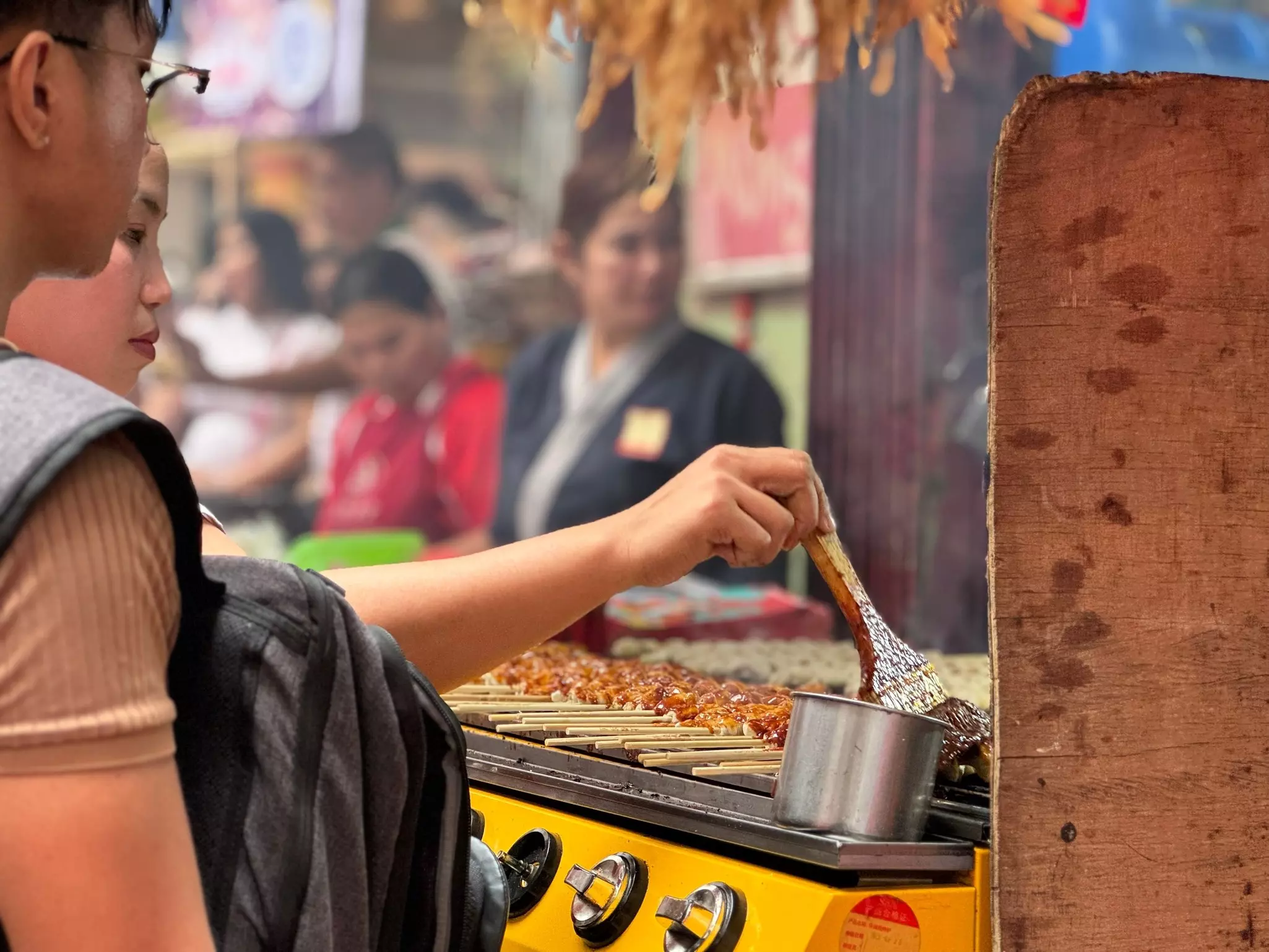 Woman cooking vegan skewer at an outdoor street food stall on a busy street.