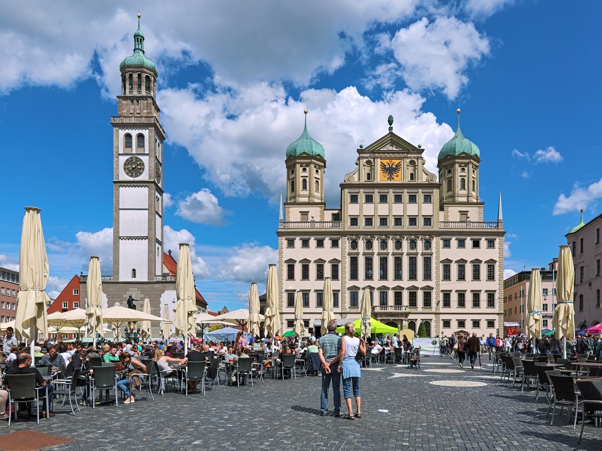 A busy street of pedestrians sitting and walking in front of the Augsburg Town Hall and Perlachturm bell tower in Augsburg, Germany.