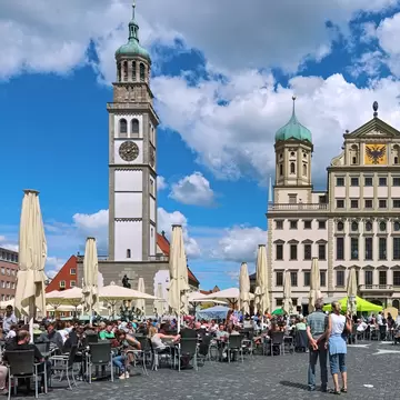 A busy street of pedestrians sitting and walking in front of the Augsburg Town Hall and Perlachturm bell tower in Augsburg, Germany.