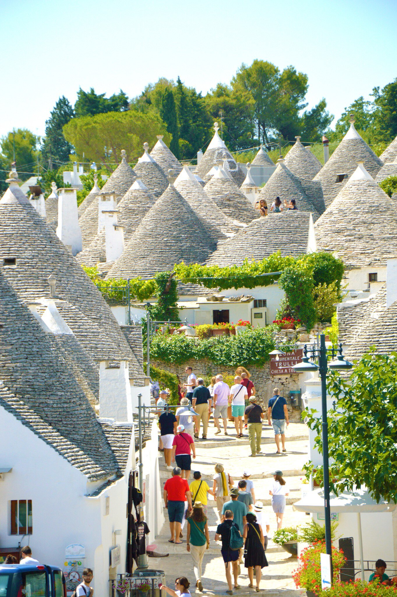 Crowd of people walking up a paved lane between whitewashed trulli houses with grey conical stone roofs in Alberobello.