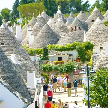Crowd of people walking up a paved lane between whitewashed trulli houses with grey conical stone roofs in Alberobello.