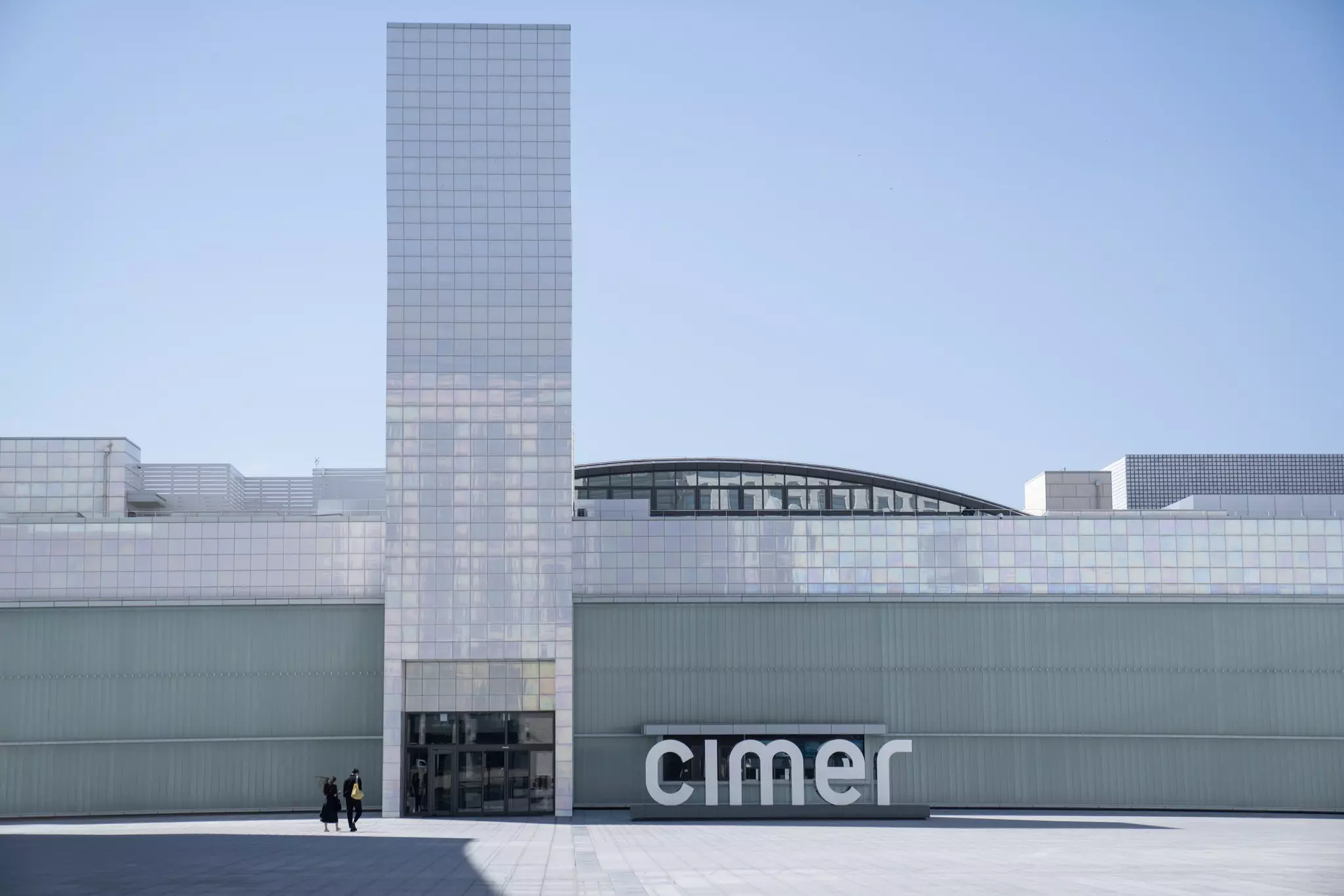 Two people walk towards a vast tiled building with a huge central rectangular tower and a sign that reads "Cimer."