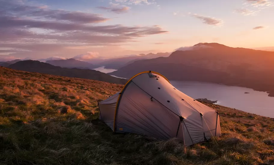 Tent pitched on a grassy hillside above Loch Lomond at sunset, with wooded islands and layered hills fading into pink sky