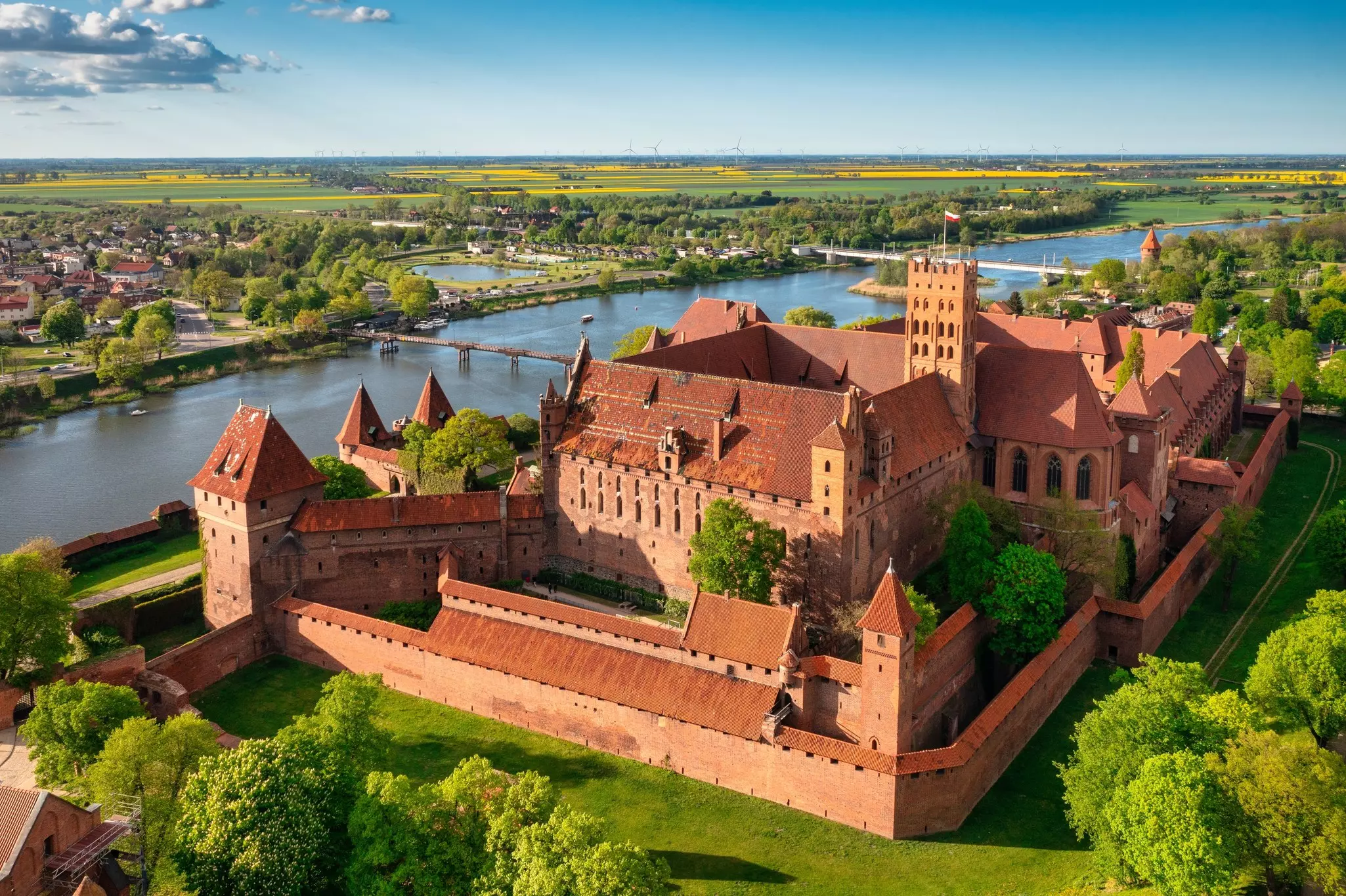 An aerial view of a monumental castle made from brick, on the bank of a river.