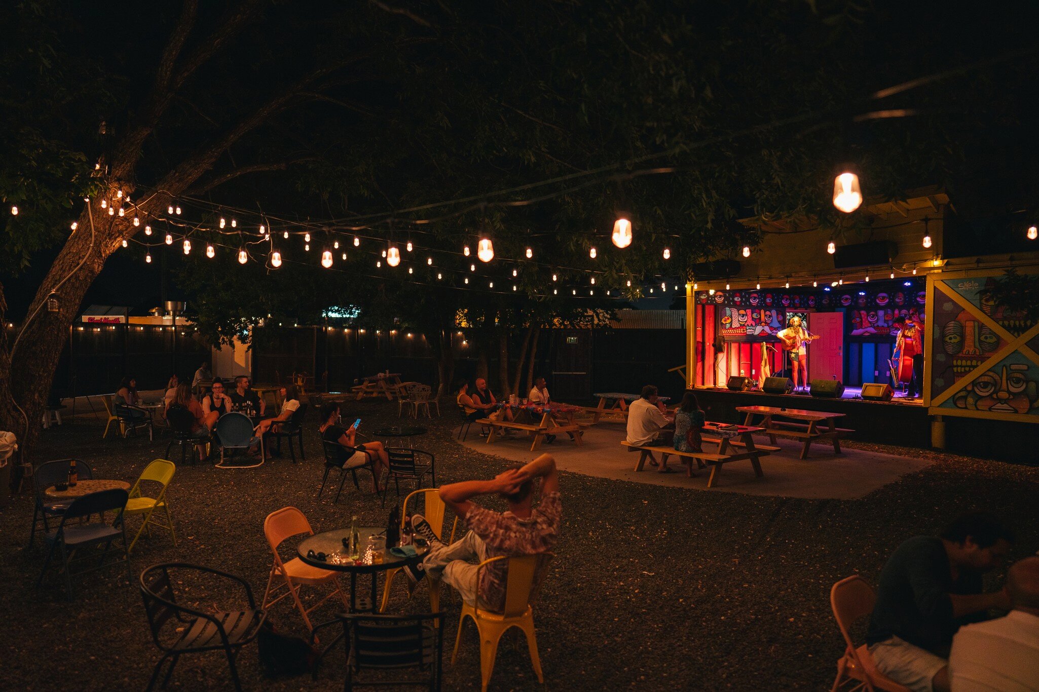 A band plays on a stage at an outdoor venue; people are sitting at tables, and lights are strung overhead.