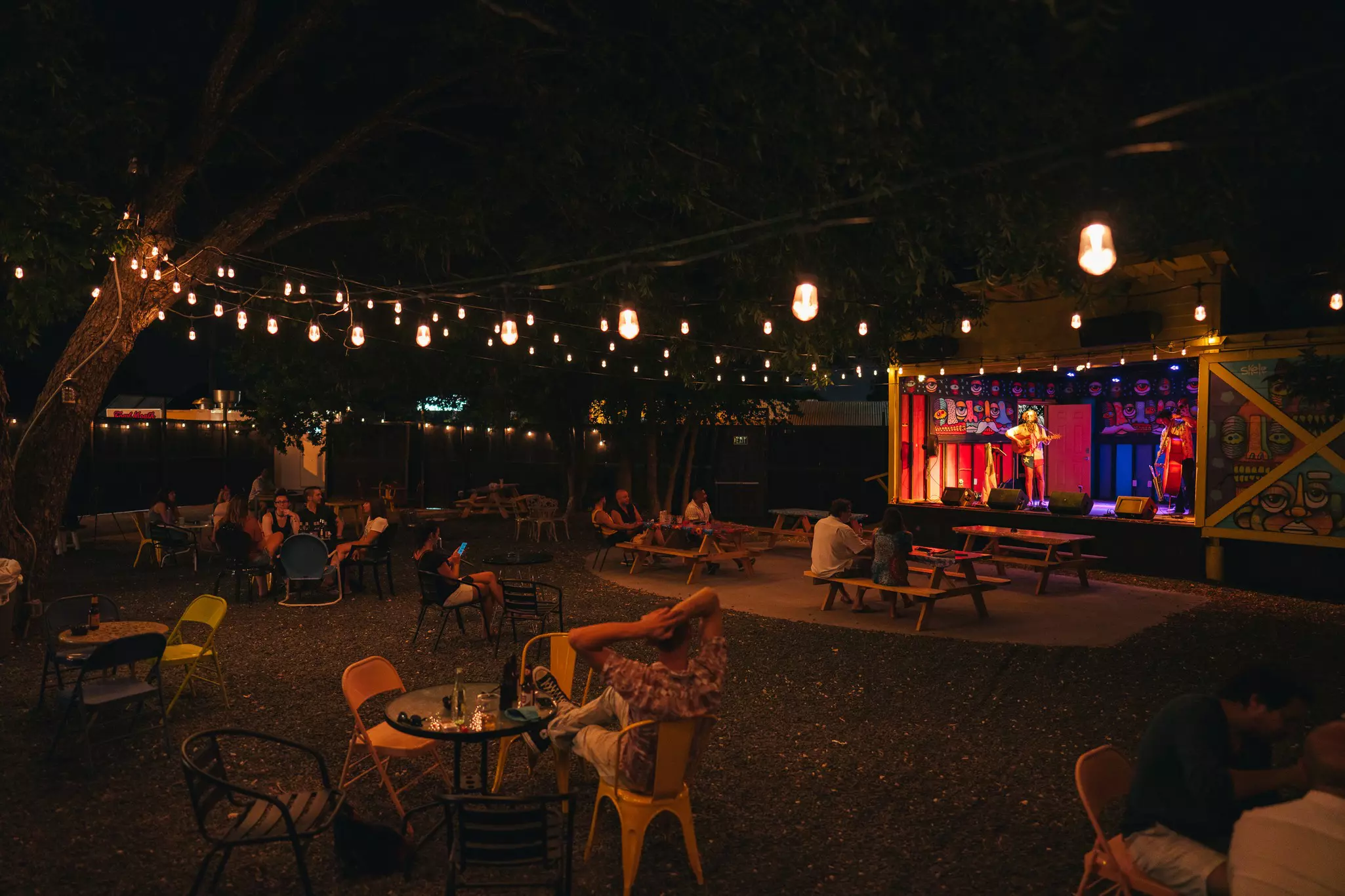 A band plays on a stage at an outdoor venue with people relaxing at tables. Twinkle lights are strung overhead