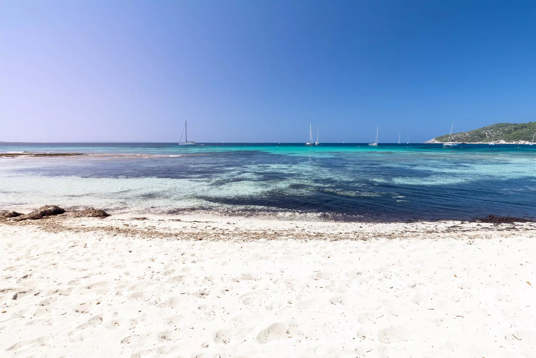 A view of an empty white-sand beach. Sailboats are visible in the azure waters of a cove beyond the beach.