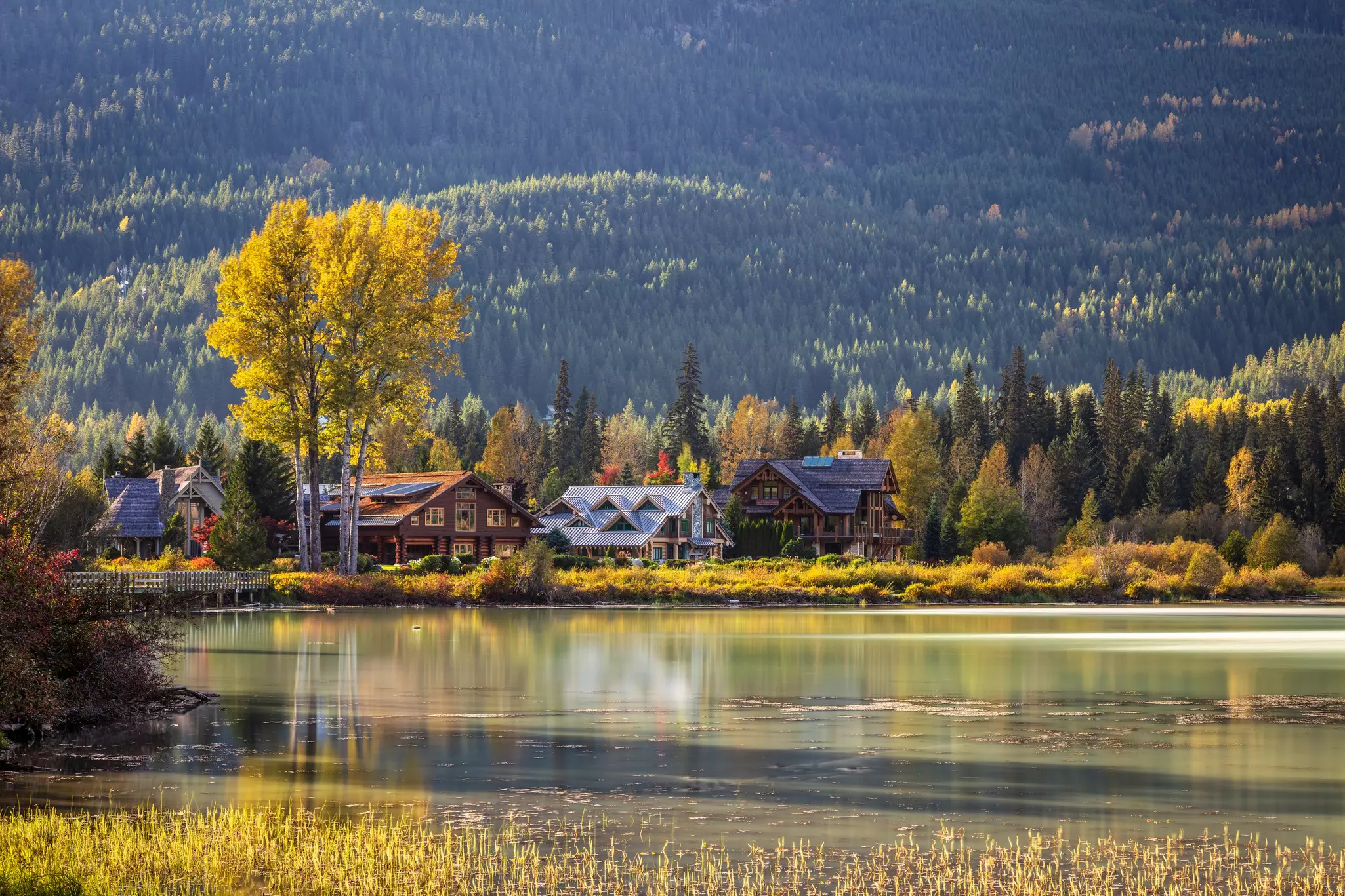 A series of large chalet-style houses line a lake. The leaves on the surrounding trees are changing to yellow in the autumn sunshine.