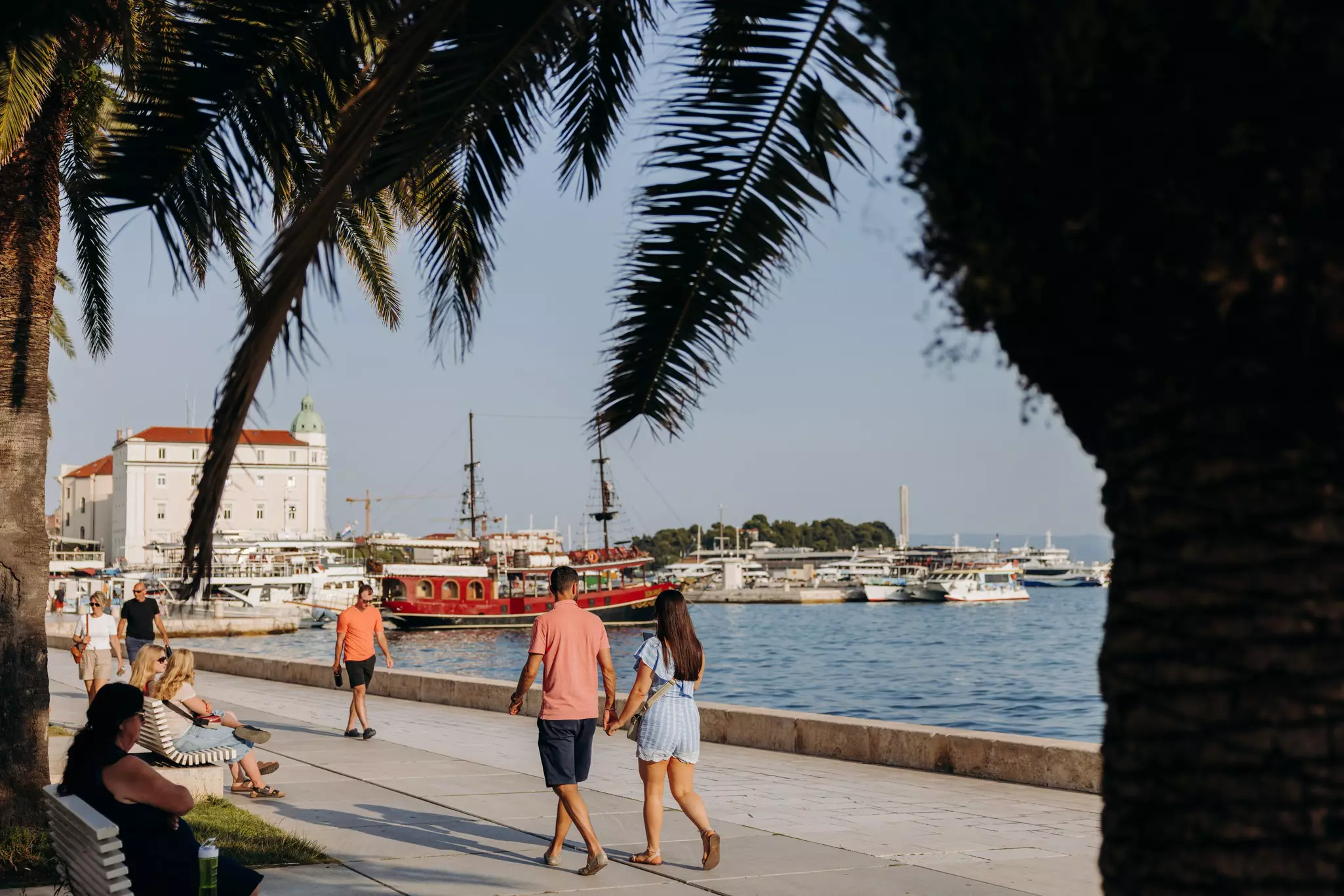 People walk along a waterfront in sunshine.