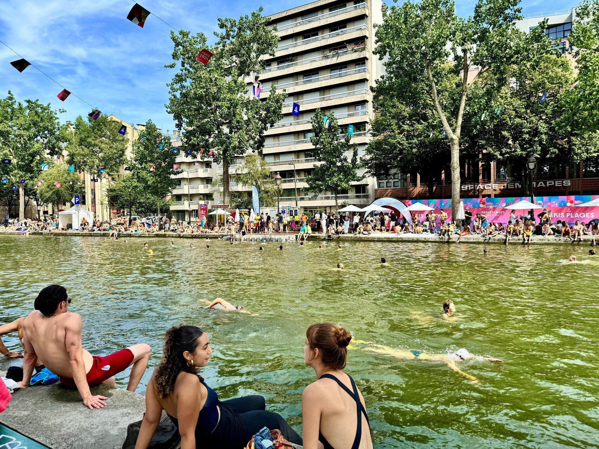 People in bathing suits sit along the edge of a city canal that is set up for summer recreation. Other people can be seen swimming in the water.