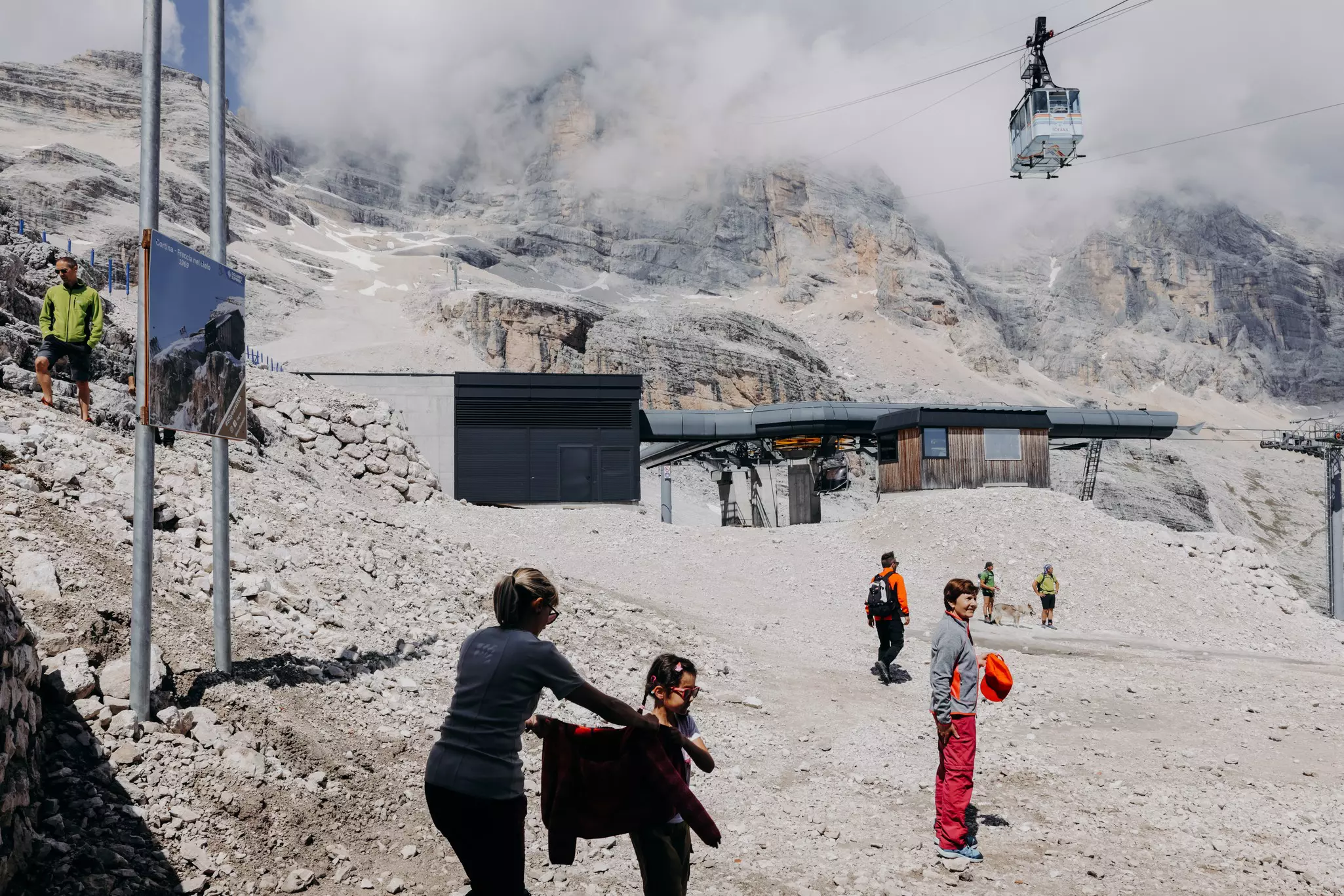 A family and other hikers walk on a rock-covered trail of a ski slope in the warmer months. A gondola is seen overhead ascending to cloud-covered mountains.