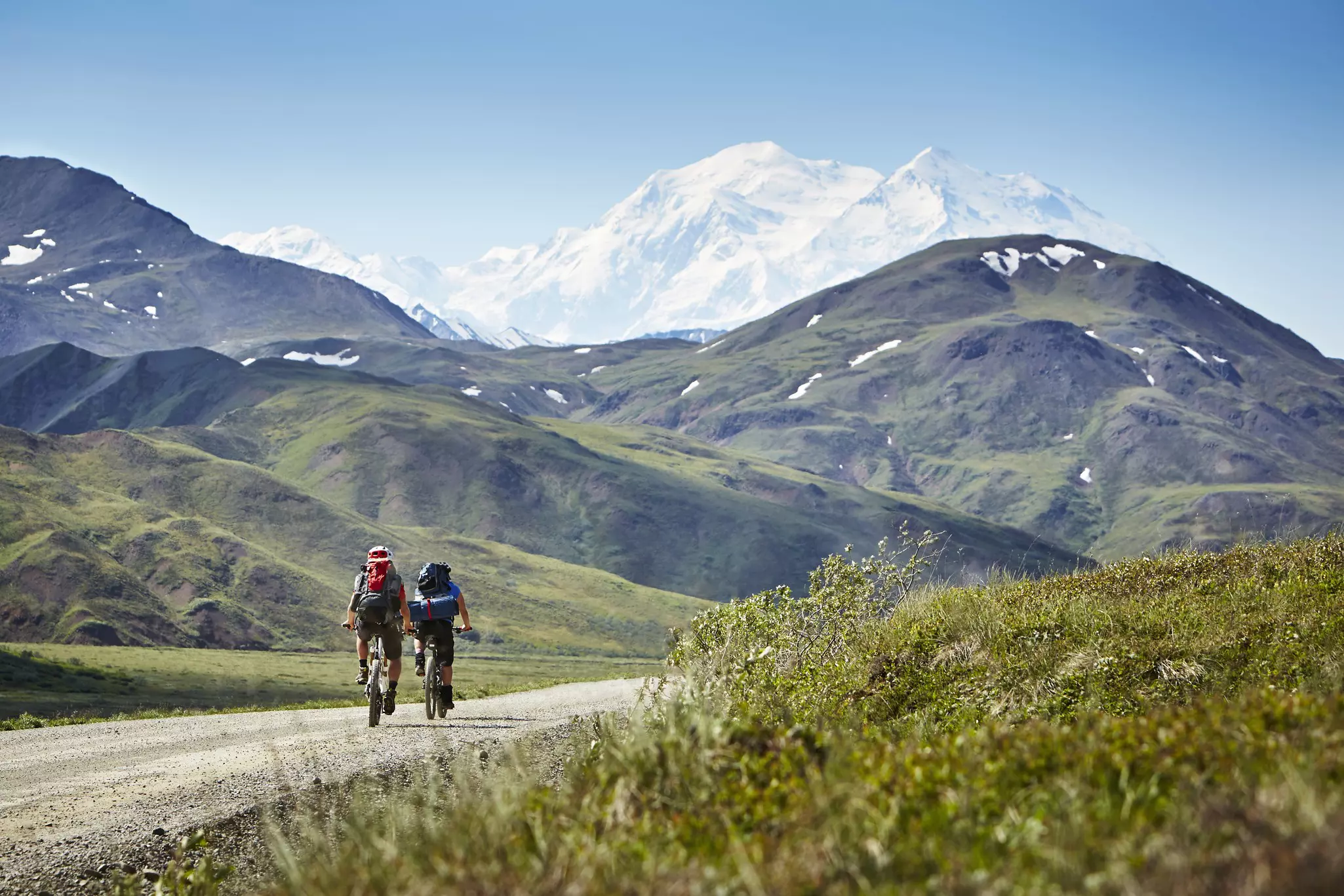 Two cyclists ride down a gravel track in a mountainous landscape.