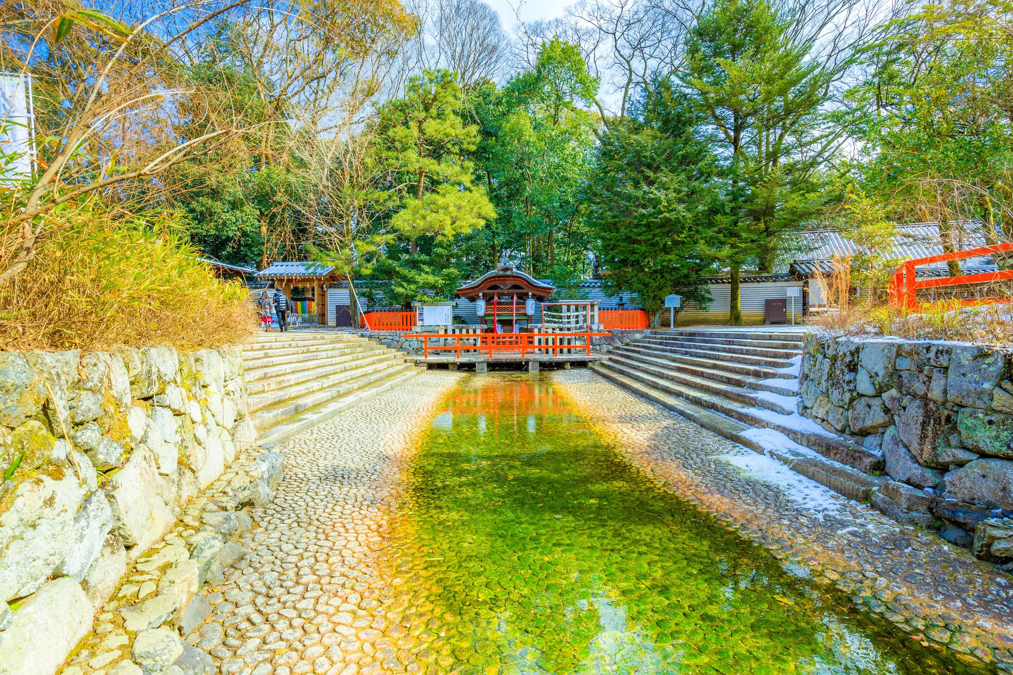 A narrow waterway leads to a small shrine protected by red gates and surrounded by trees.