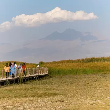 Women walking a wooden boardwalk through golden reeds, with the hazy twin peaks of Mount Erciyes rising beyond