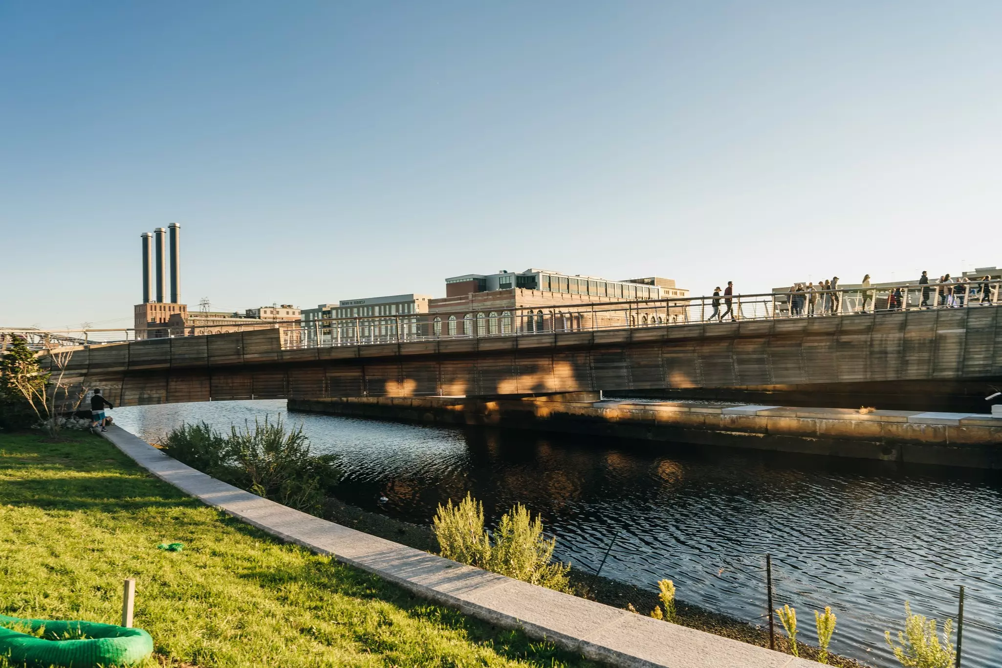 Wide shot of cement walkway along a river with a cement bridge above and pedestrians on it, with low-rise city buildings in the background.