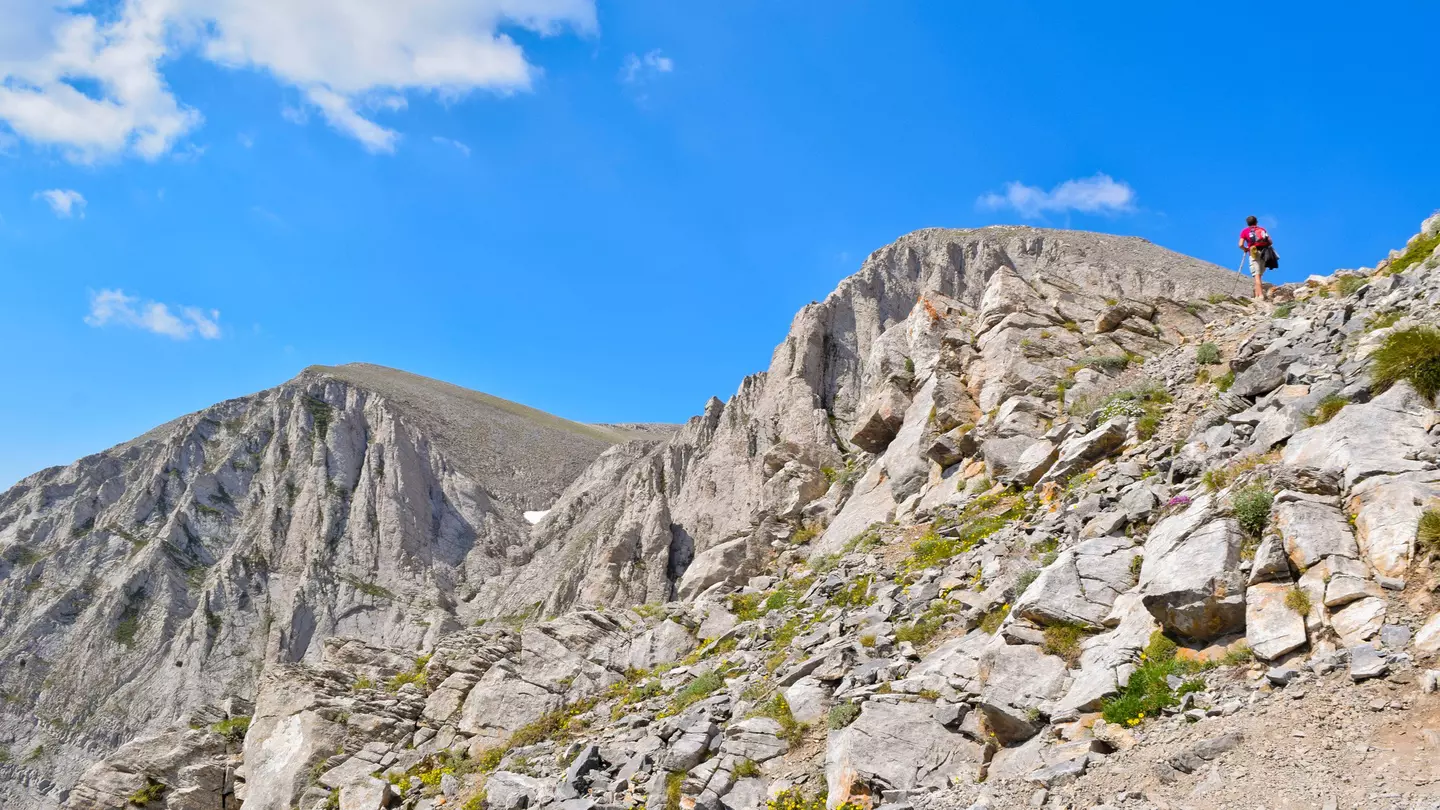 A hiker walks up a steep, rocky path to the peak of Mt Olympus. The mountain peak is bare and rocky and a blue sky is visible above.