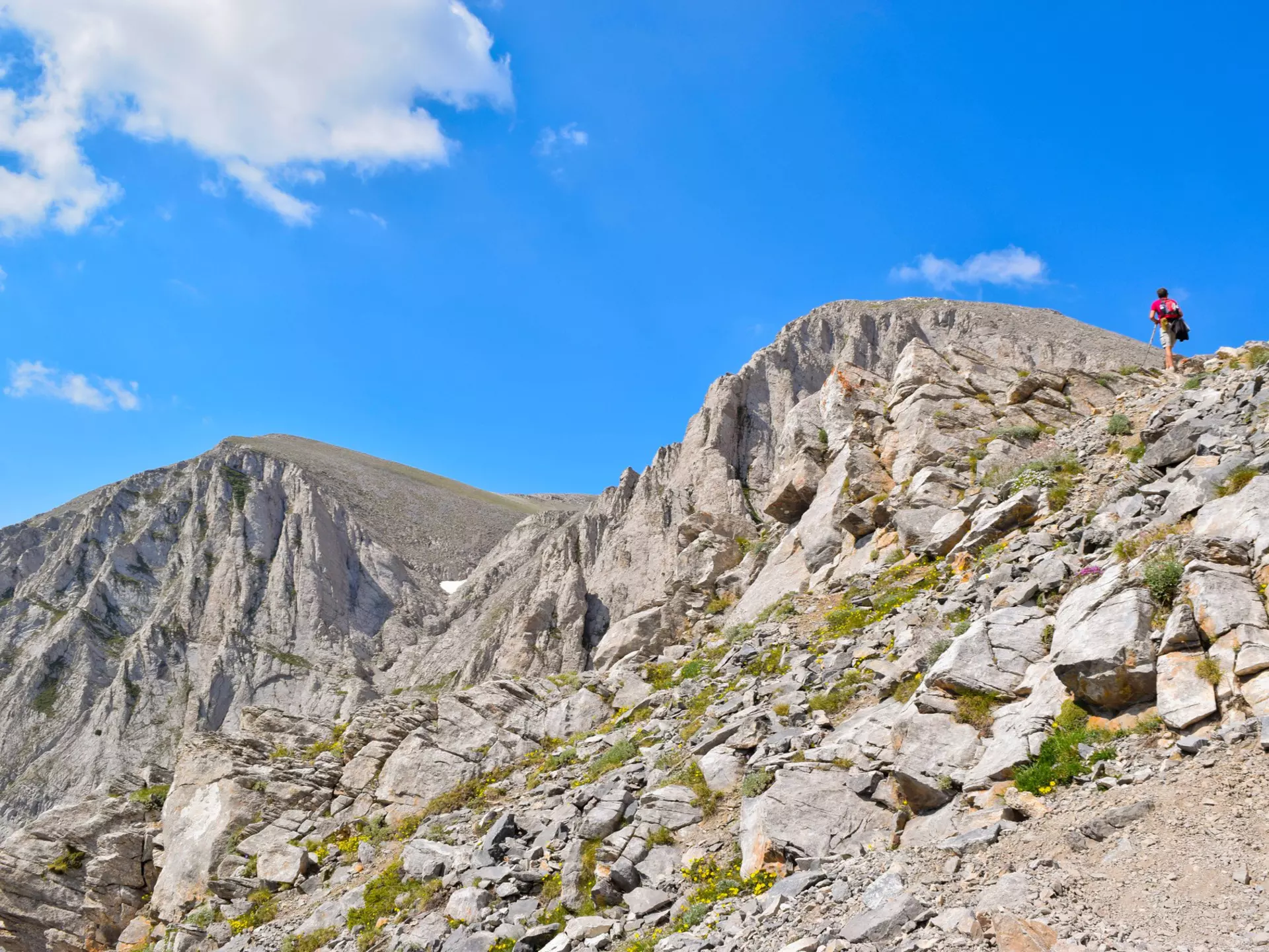 A hiker walks up a steep, rocky path to the peak of Mt Olympus. The mountain peak is bare and rocky and a blue sky is visible above.