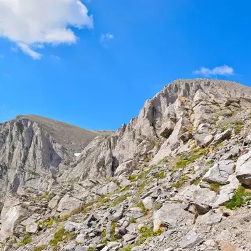 A hiker walks up a steep, rocky path to the peak of Mt Olympus. The mountain peak is bare and rocky and a blue sky is visible above.
