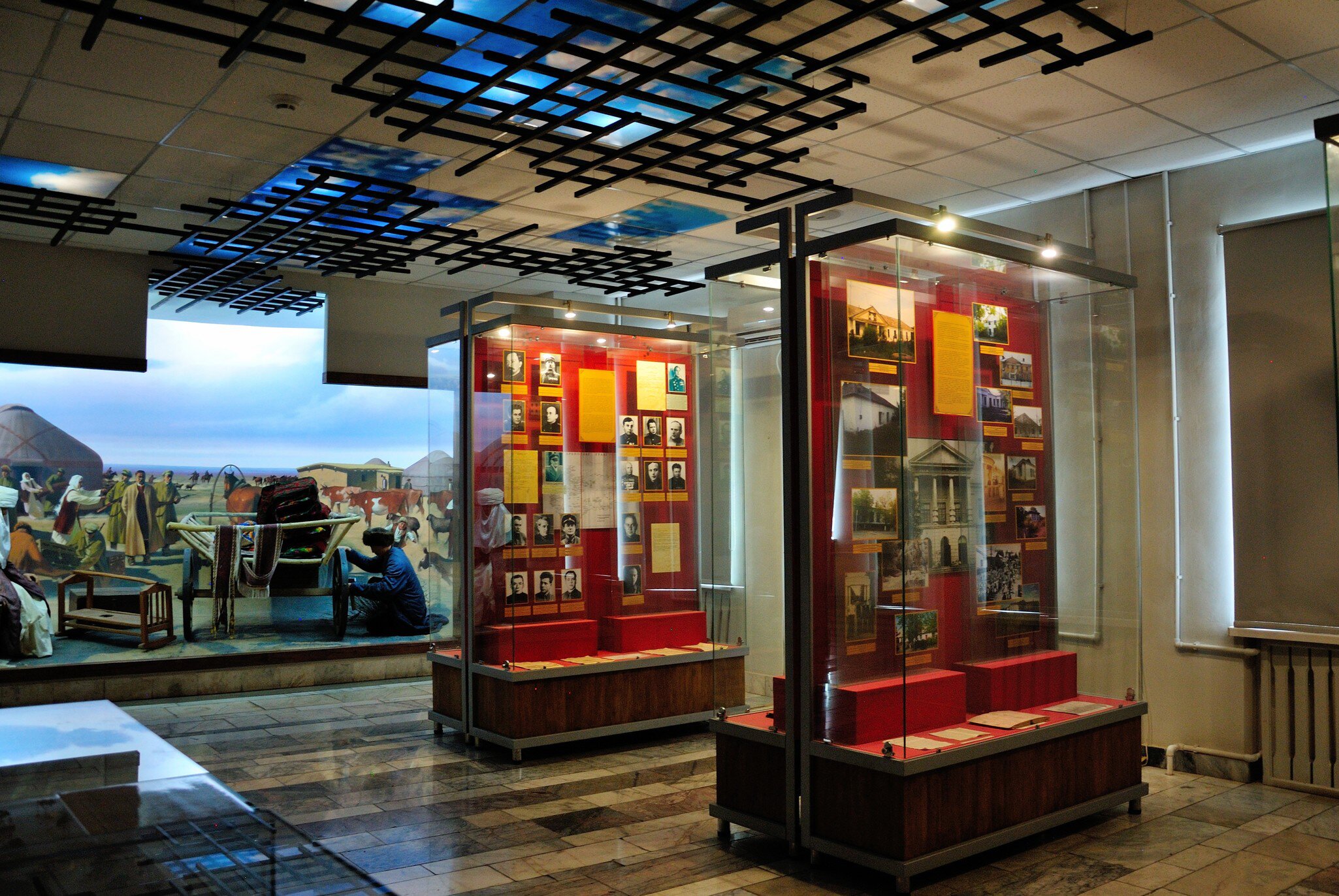 Two lit glass cases of prisoner photographs and documents on red backing boards, under a ceiling grid of black bars, beside a painted diorama of a steppe camp.
