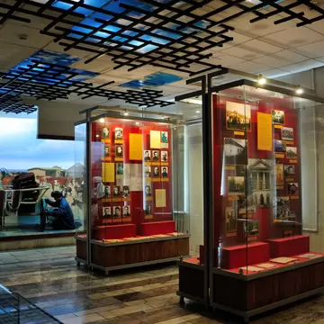 Two lit glass cases of prisoner photographs and documents on red backing boards, under a ceiling grid of black bars, beside a painted diorama of a steppe camp.