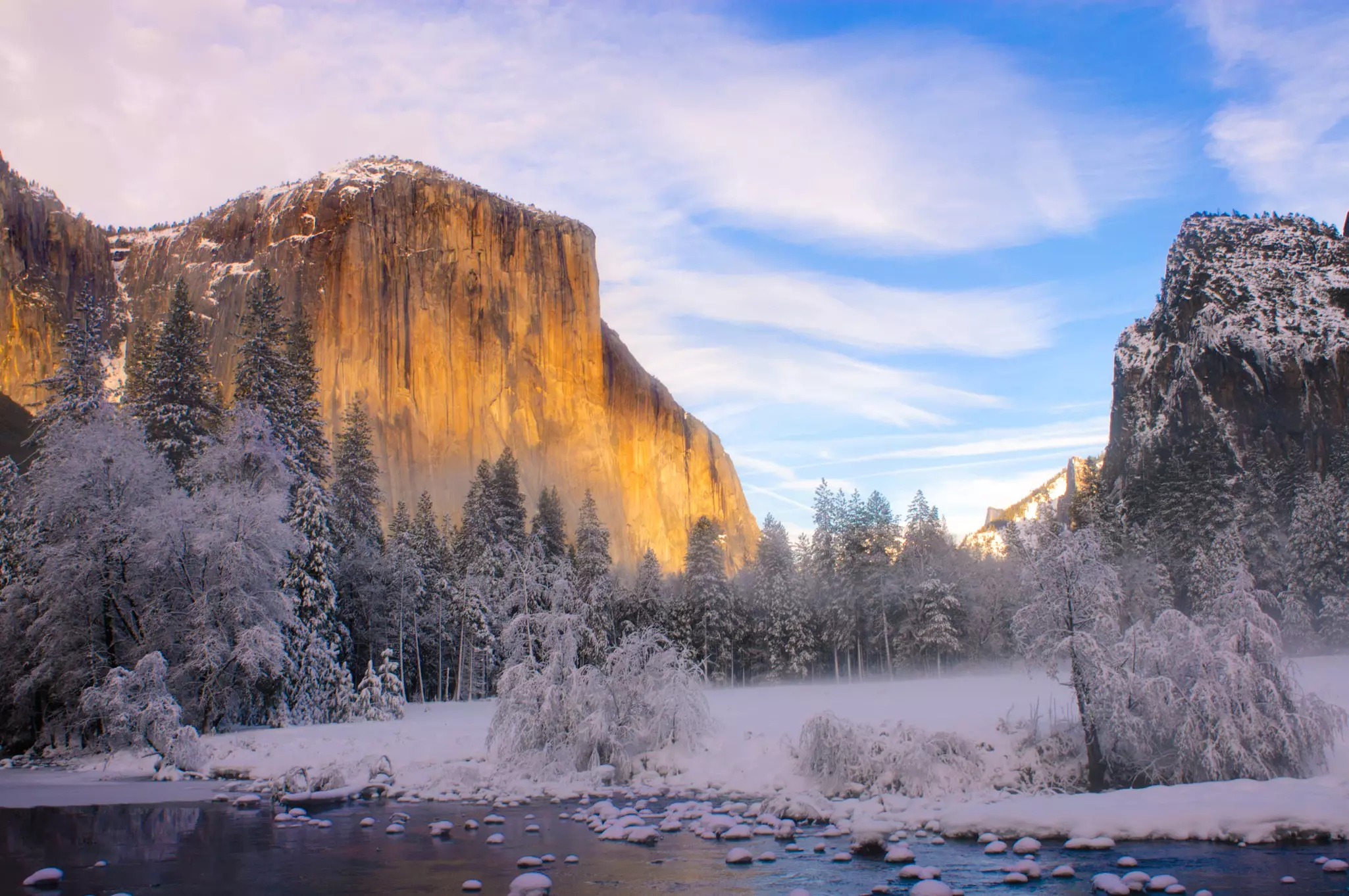 A rock face in streaks of sunlight towers above a snowy landscape in shade, with a river in the foreground.