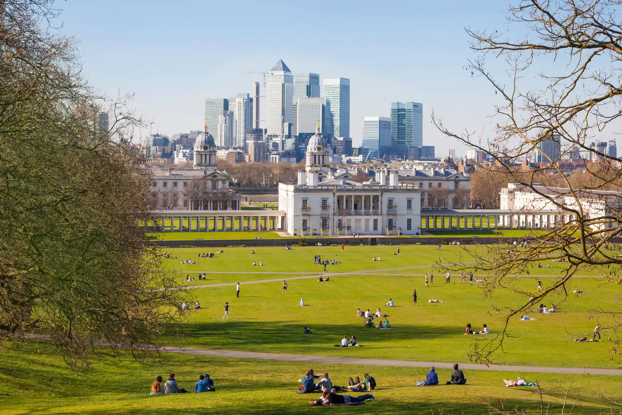 People enjoying parkland on a sunny day. The view stretches to a symmetrical old naval building with modern skyscrapers in the distance; Greenwich, London