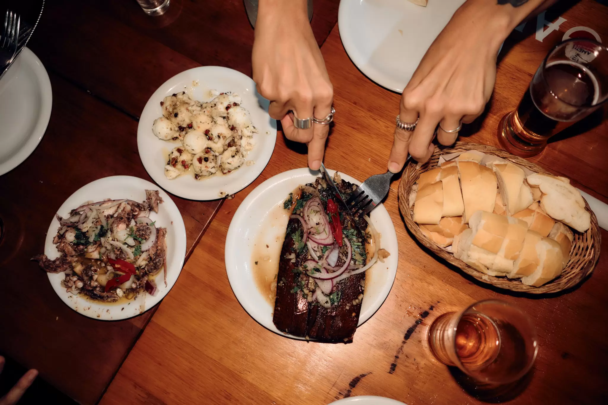 Hands using a fork and knife to cut into a meal on a plate; other dishes and glasses of beer are also on the wooden table.