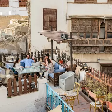 Friends chat on a rooftop terrace beside carved wooden roshan balconies of old coral-stone houses in Al-Balad, Jeddah