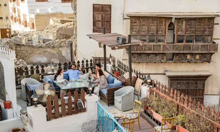 Friends chat on a rooftop terrace beside carved wooden roshan balconies of old coral-stone houses in Al-Balad, Jeddah