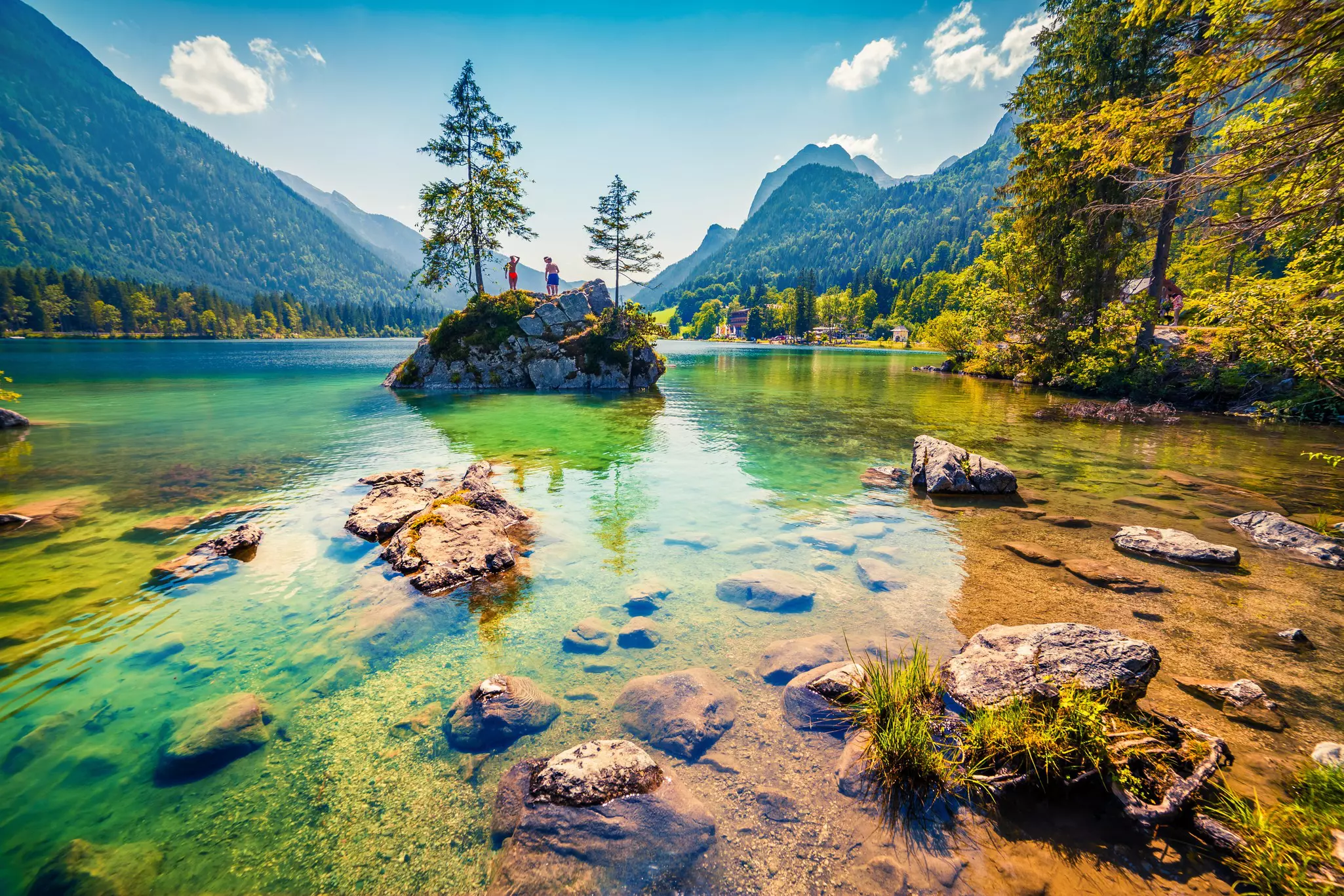 Two people stand on a rocky island in a beautiful, shallow lake surrounded by mountains.