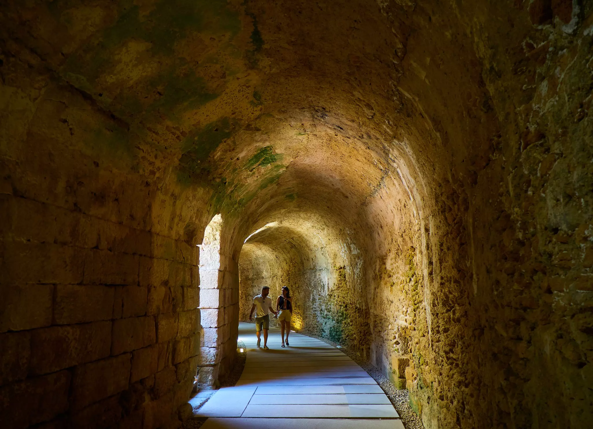 Two people walk down a curving corridor underground within an ancient structure.