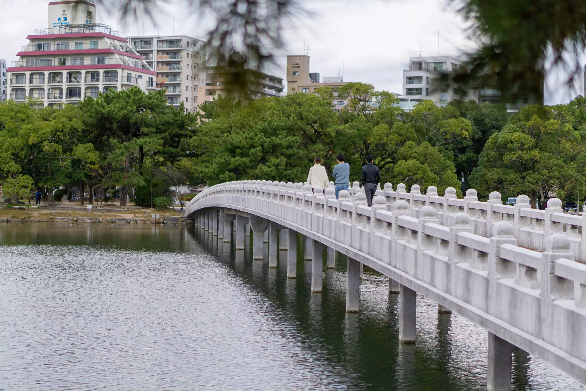 Three people walking across a bridge in a park. A body of water flows underneath the bridge.