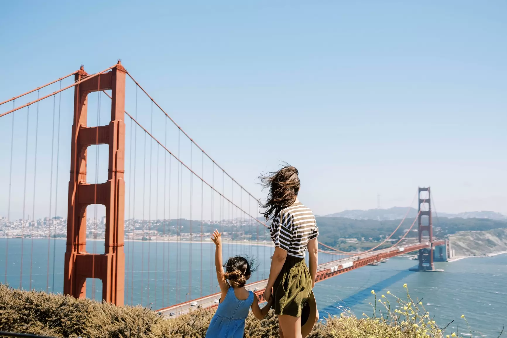 A woman and young girl hold hands overlooking San Francisco's Golden Gate Bridge on a clear day