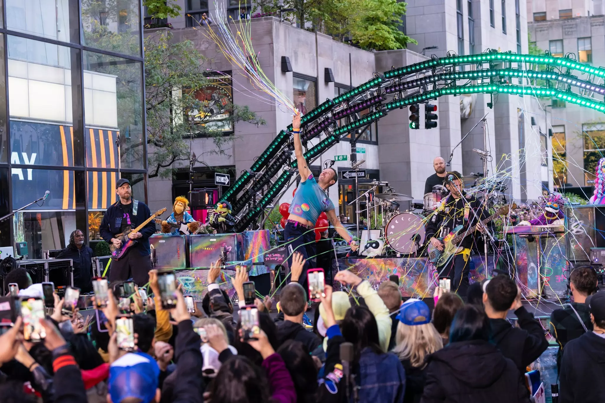A band performs in front of a live audience during a taping in a city plaza.