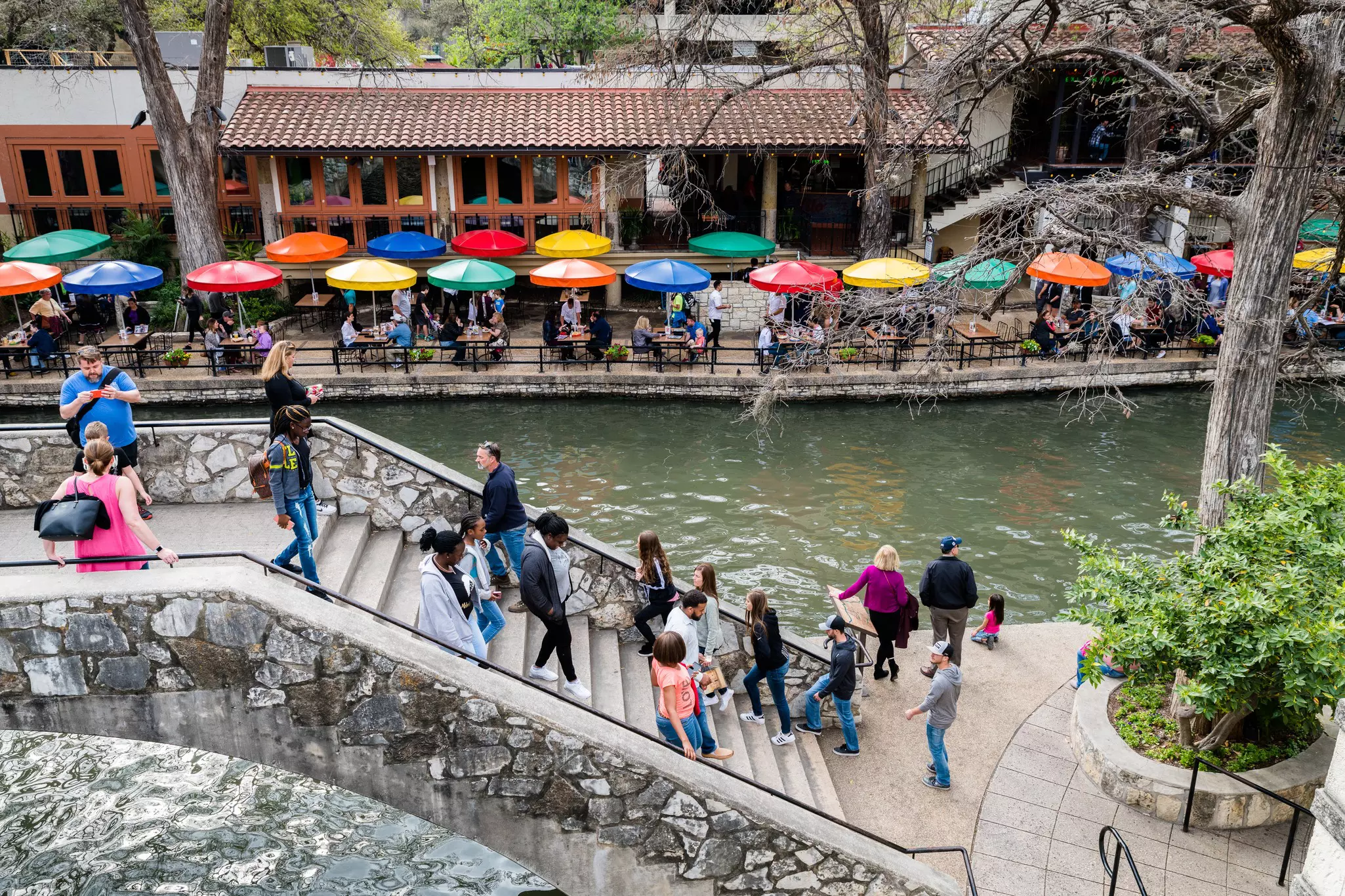 People on stairs by the river in San Antonio, Texas; on the other shore, people sit at tables with brightly colored umbrellas.