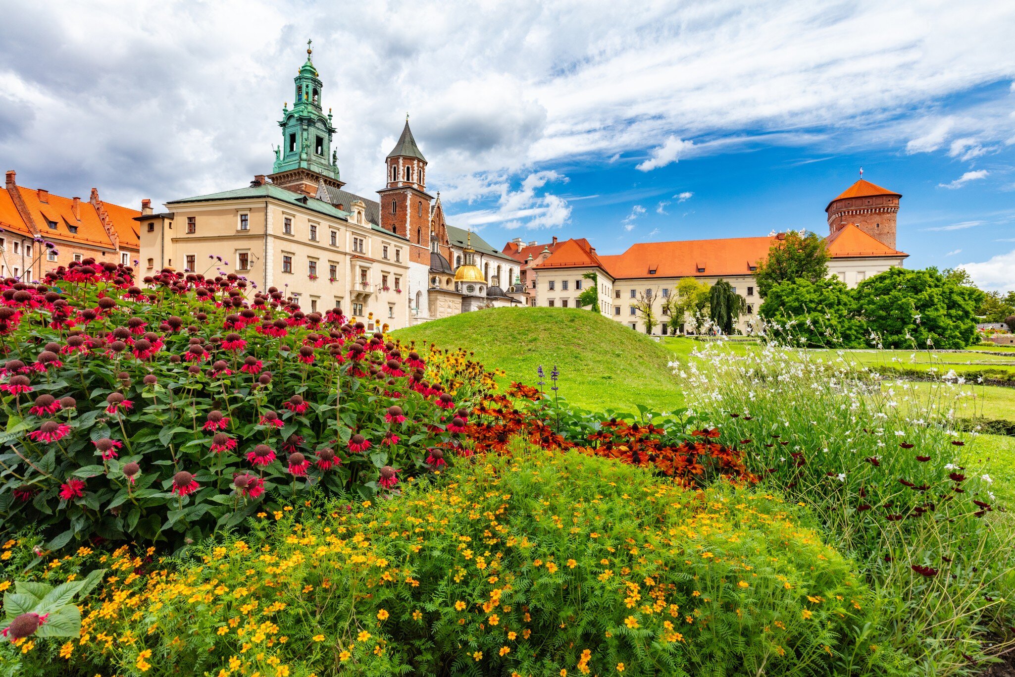 Colorful flower beds in front of a castle's towers and terracotta roofs.
