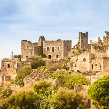 Crumbling stone tower houses on a scrubby hillside at Vathia in the Mani, Greece, with pine trees below and sea haze behind.