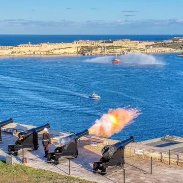 A cannon fires at noon from the Saluting Battery in Valletta, Malta, with Birgu in the background.