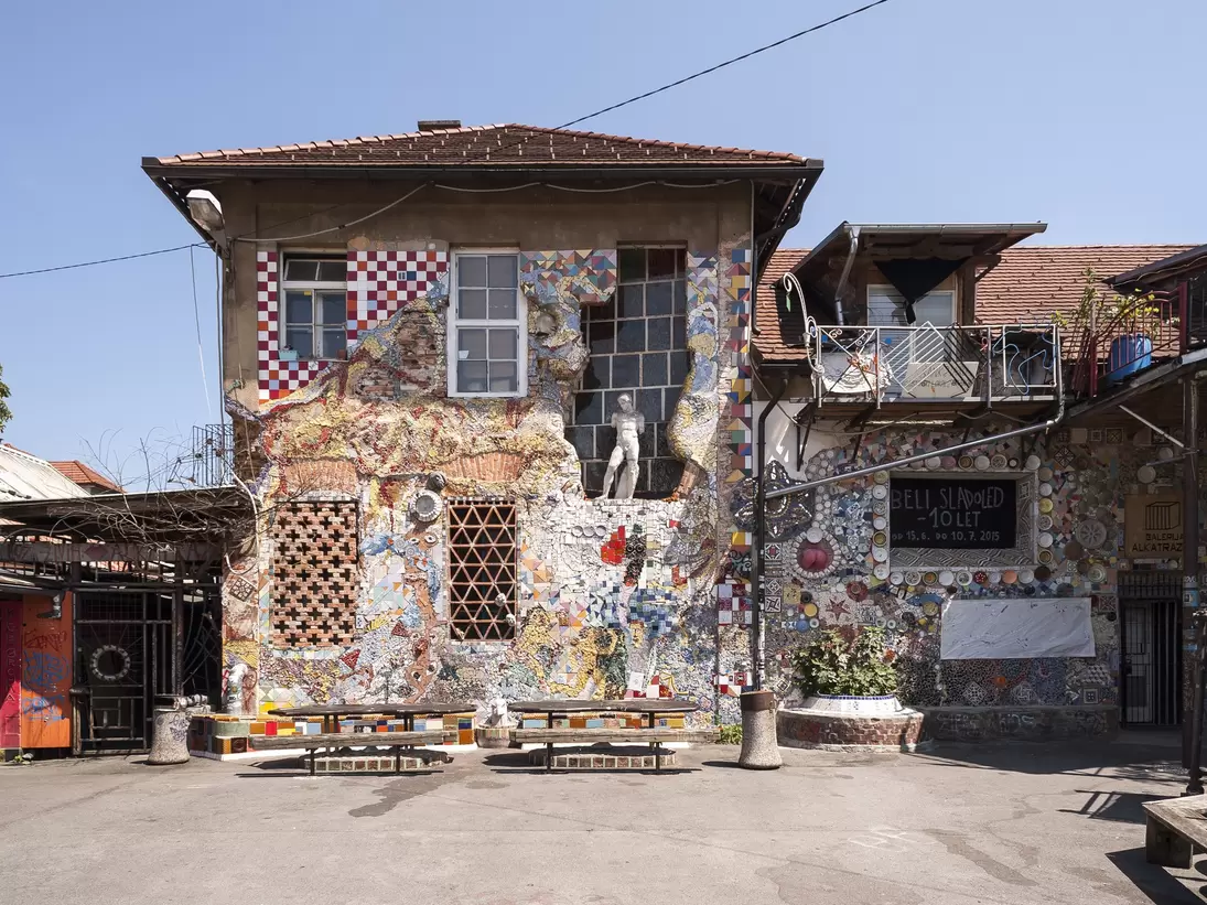 Metelkova building covered in tile mosaics with a white statue in a window, picnic tables and signs for Galerija Alkatraz