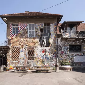 Metelkova building covered in tile mosaics with a white statue in a window, picnic tables and signs for Galerija Alkatraz
