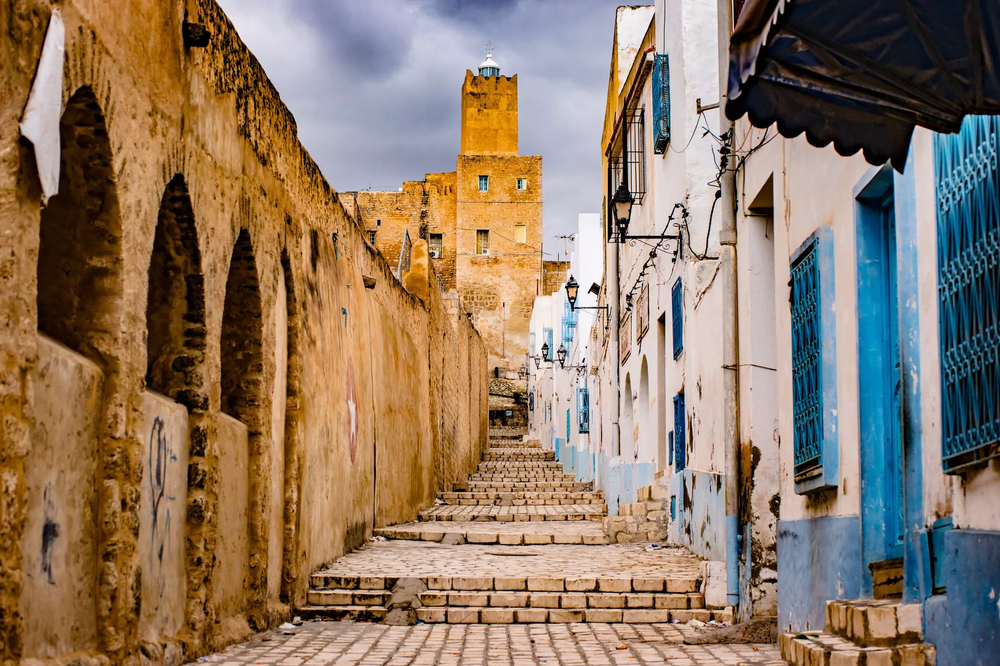 Blue and white buildings line a brick pathway with stairs leading up to a tower.