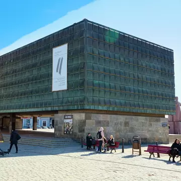 Dark copper-clad museum block raised on stone pillars, with people on benches in a cobbled square, Rīga