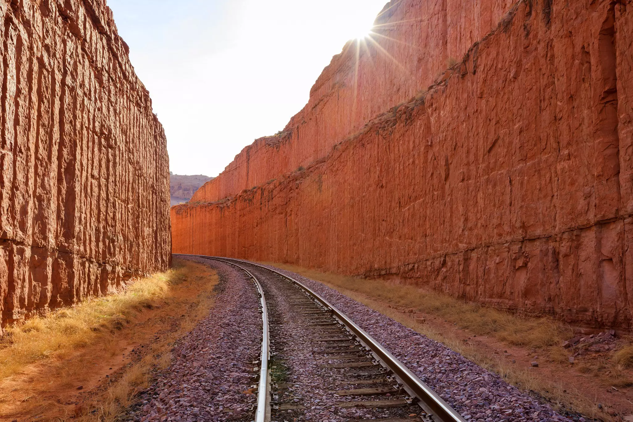 Railway tracks carve their way through a rocky canyon with sheer red-rock walls rising above the train line.
