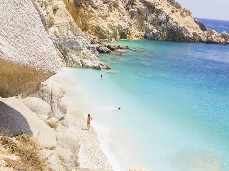 A woman stands on the shore of a white beach, with clear blue ocean. 