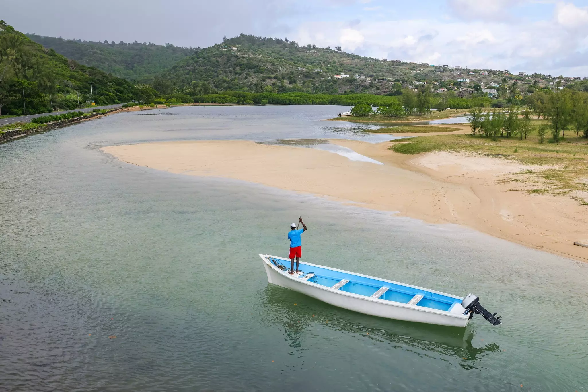 An overhead view of a fisherman standing on a small wooden boat in a shallow inlet by a beach on a tropical island.