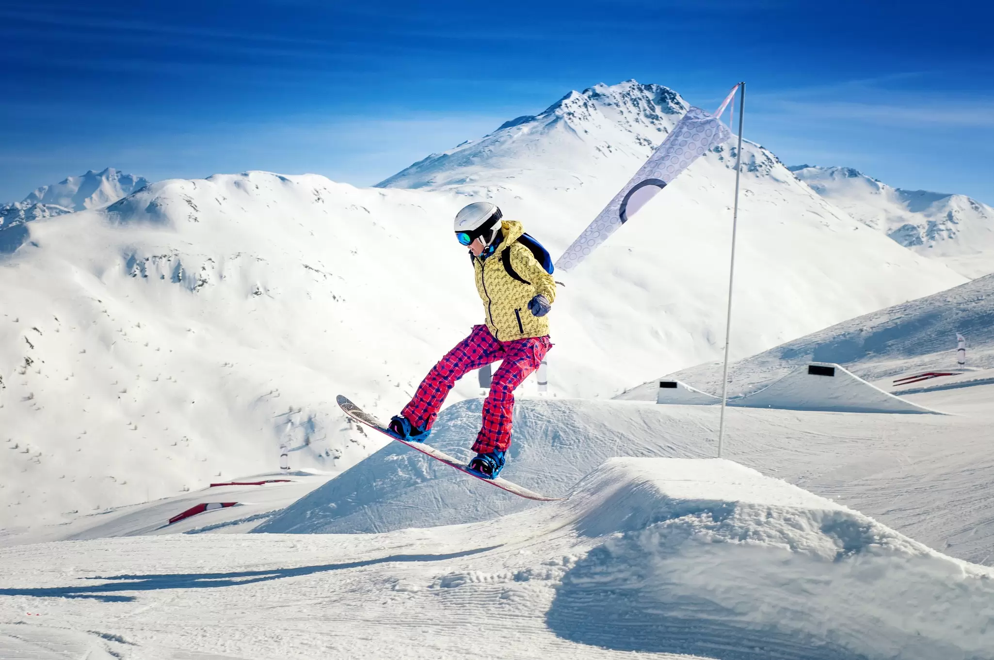 A snowboarder in a yellow jacket and pink pants is slightly airborne over snow.
