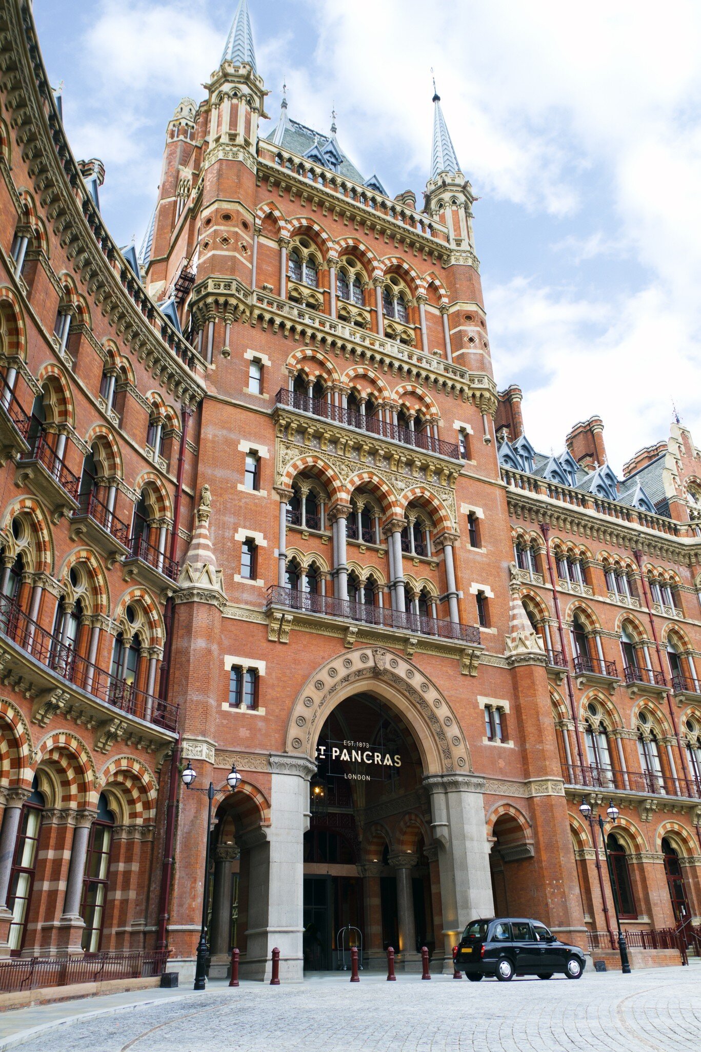 A black taxi stands outside a large Gothic-style red-brick building with many windows.
