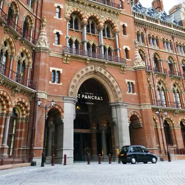 A black taxi stands outside a large Gothic-style red-brick building with many windows.